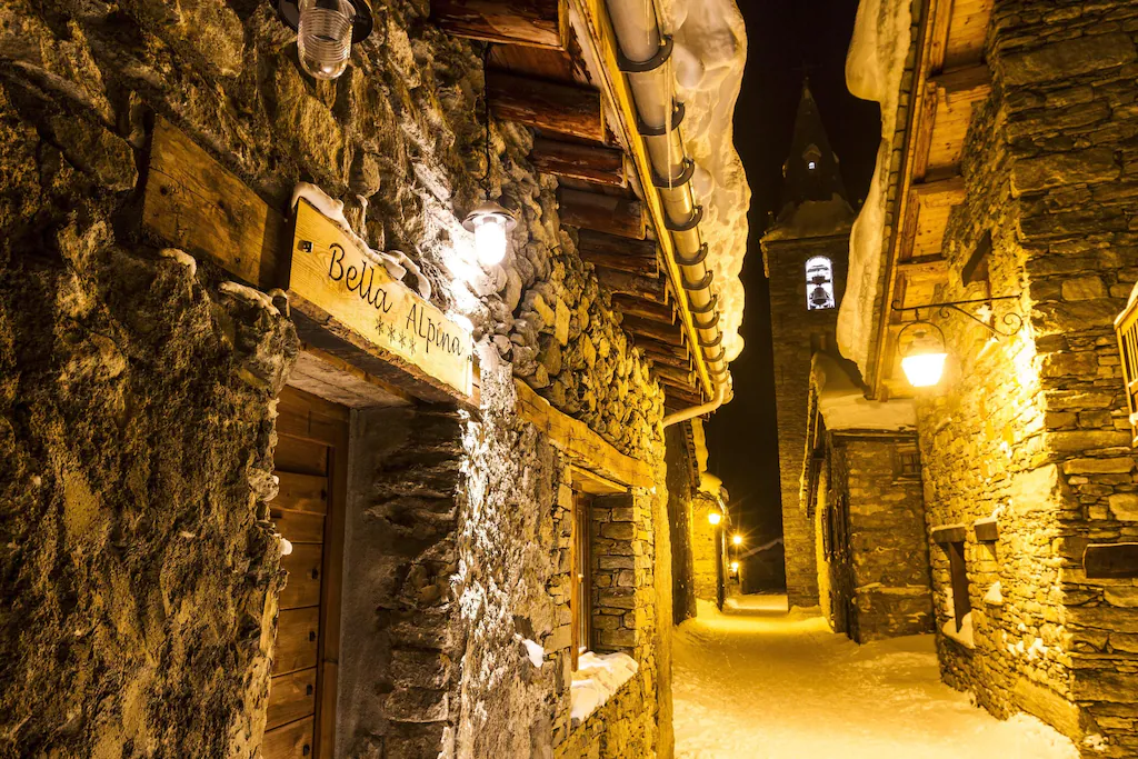 Façade du chalet Bella Alpina la nuit, ruelle enneigée et clocher de Bonneval-sur-Arc